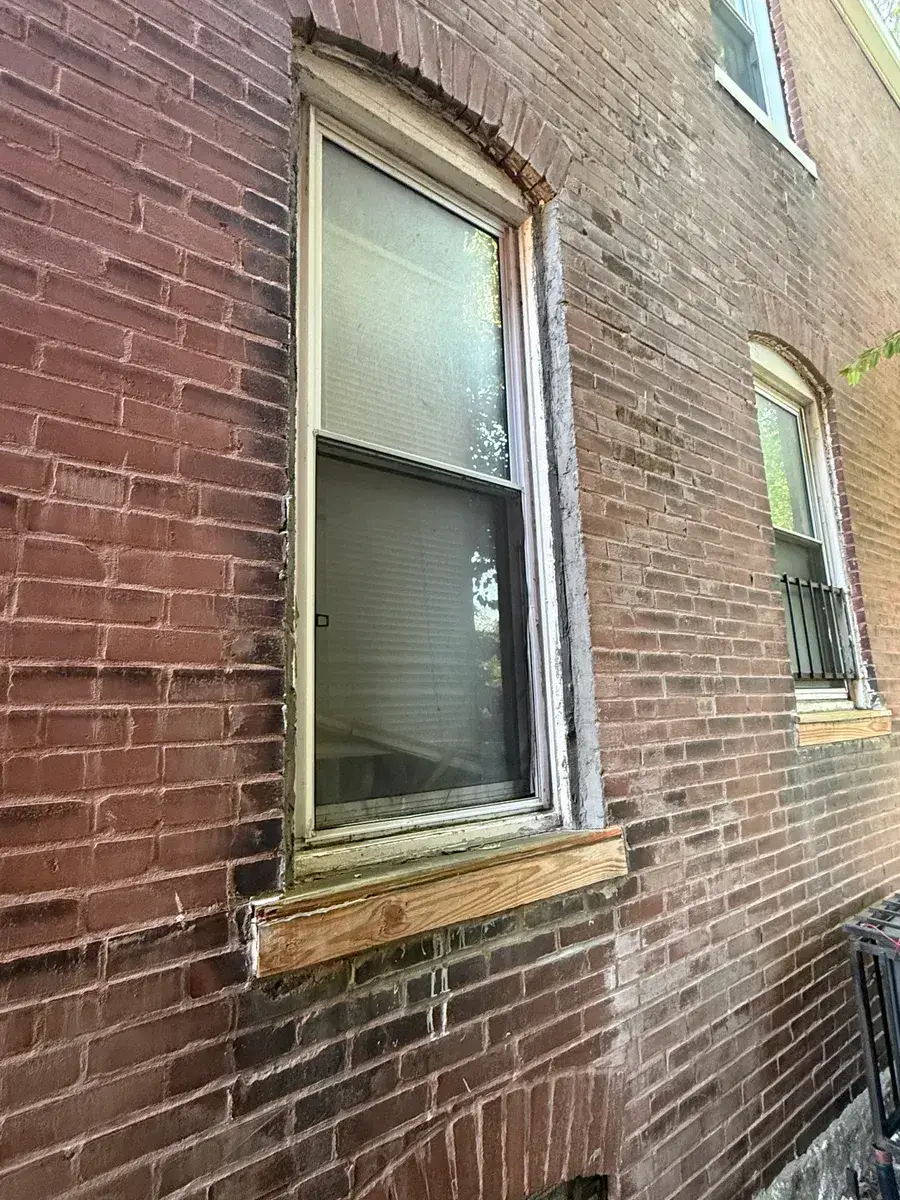 Old window with weathered, deteriorating wood trim set in an arched brick opening before replacement